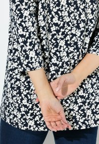 Námořnická modrá blůza s bílým květinovým vzorem, tříčtvrteční rukávy a zakřivený lem. Ruce sepnuté za zády.