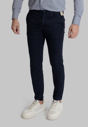 Uomo che indossa pantaloni slim fit blu navy, camicia azzurro chiaro con bottoni e sneakers casual bianche, in piedi su uno sfondo grigio.