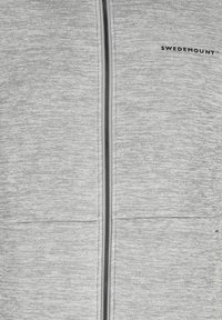 Graue Zip-Jacke aus strukturiertem Stoff mit einem zentralen Reißverschluss und zwei vorderen Taschen. Logo auf der oberen linken Seite angebracht.