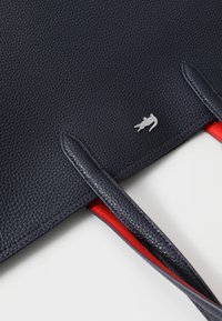Sac fourre-tout en cuir marine avec une finition texturée, doté d'une quincaillerie en logo argenté et d'une doublure intérieure rouge. Longs anses plates pour le transport.