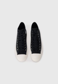 Sneakers alta in canvas nero con punte in gomma bianca e occhielli dorati, dotate di lacci piatti neri e una texture liscia.