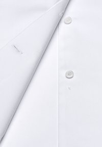 Chemise blanche avec un col pointu, patte de boutonnage et texture tissée simple. Présente un petit accroc près du col.