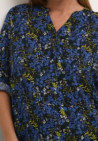 Blumenbluse mit dunkelblauem Hintergrund, mit blauen und weißen Blumen, grünen Blättern, einem V-Ausschnitt und einem Knopfdetail am Kragen.
