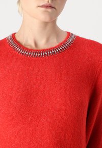 Pull rouge avec un col rond orné d'une rangée d'embellissements argentés, en maille texturée, et avec des manches longues.
