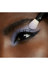 Nærbilde av et mørk hudtone øye med myk lavendeløyenskygge, kraftig svart eyeliner og en børste som påfører sminke nær brynet.
