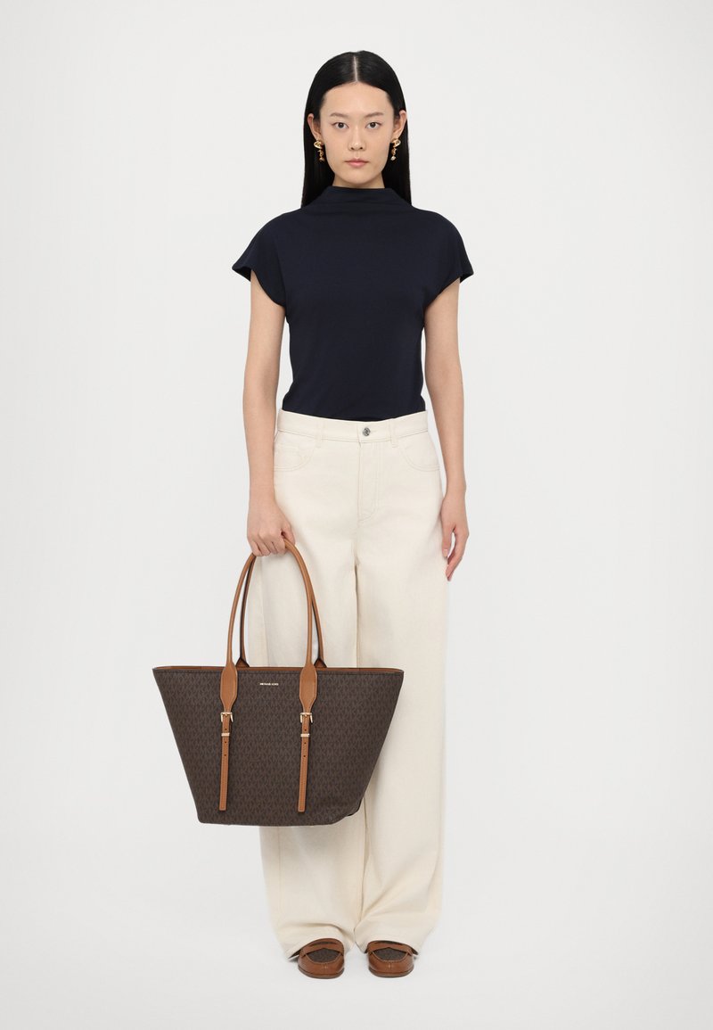 Brun tote taske med lysebrune læderhåndtag, prydet med et logo-mønster. Båret af en person i en marineblå bluse og cremefarvede vide bukser.