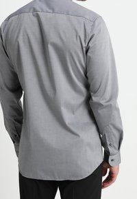 Szara koszula z długim rękawem o gładkiej fakturze, z delikatnym połyskiem, zaokrąglonym dołem oraz mankietami zapinanymi na guziki. Widok z tyłu ukazuje prosty wzór.