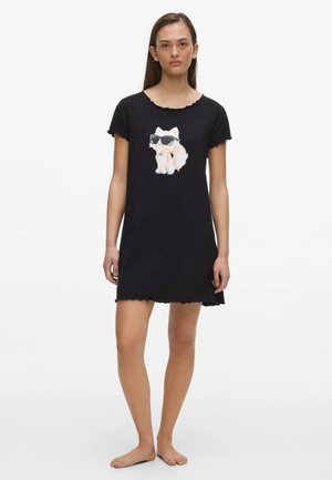 Nő fekete rövid ujjú ruhában, amin egy fehér macska látható napszemüveggel és rózsaszín masnival, mezítláb áll fehér háttér előtt.