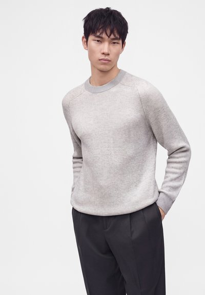 Jeune homme portant un pull côtelé gris clair et un pantalon noir, debout les mains partiellement dans les poches sur un fond uni.