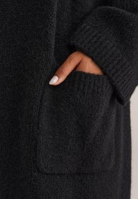 Roka ar manikīrētu nagu daļēji iekšā melnā adītā džemperī ar ribotu piedurknēm.