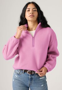 Pull polaire rose avec col montant et fermeture éclair au quart. Présente des épaules tombantes et des accents côtelés, assorti à un jean bleu déchiré.