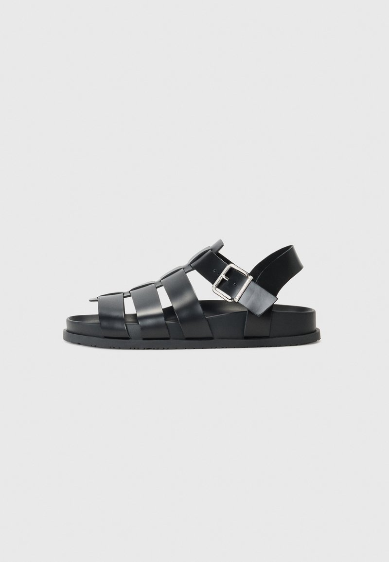 Sandálias de couro preto com um design aberto, apresentando múltiplas tiras e uma tira para o tornozelo com fivela. Sola plana com um acabamento texturizado.