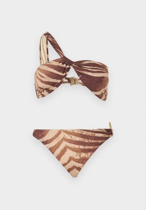 Femme debout pieds nus portant un bikini asymétrique imprimé zèbre marron et beige, ainsi que des lunettes de soleil teintées ambrées, sur un fond uni.