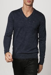 Homme portant un pull à col en V à manches longues bleu foncé et un pantalon noir, marchant devant un fond gris clair uni.