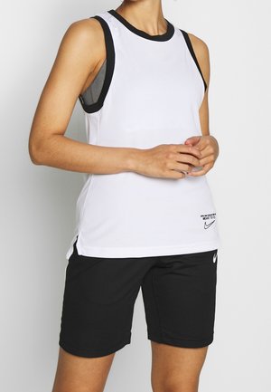 Persona che indossa una canottiera bianca Nike senza maniche con rifiniture nere e pantaloncini neri Nike, in piedi con le mani intrecciate davanti.