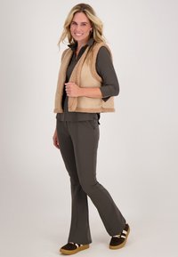 Beige fleec vest met schapenvachttextuur, gedragen over een donkergroene shirt. Uitlopende donkere broek en bruine sneakers met witte accenten maken de outfit compleet.