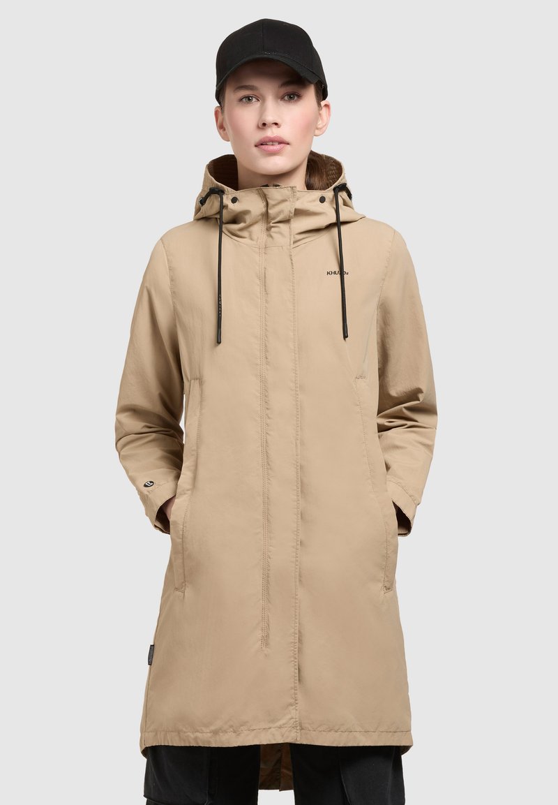 Parka impermeable beige con capucha, presenta cordones, mangas largas y bolsillos. Textura suave con corte recto y detalle del logo.