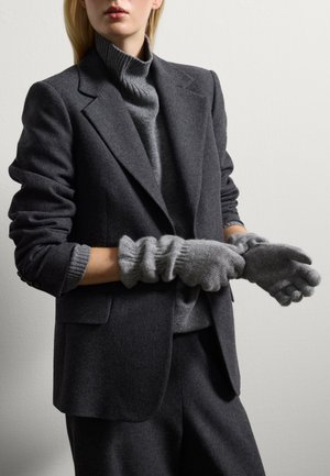 Donna che indossa un blazer e pantaloni grigio scuro, un maglione a collo alto grigio, mentre indossa dei guanti di lana grigio chiaro contro un muro chiaro e semplice.