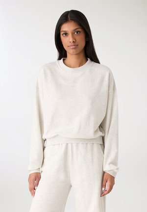 Donna con lunghi capelli scuri che indossa una felpa beige chiaro a maniche lunghe e pantaloni della tuta abbinati, in piedi davanti a uno sfondo semplice.