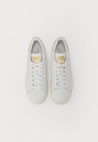 Sapatilhas brancas em couro com atacadores planos, detalhes perfurados e acentos bege no calcanhar. Etiqueta do logótipo em dourado na lingueta.