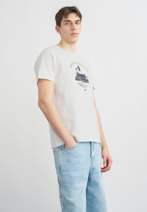 Młody mężczyzna z krótkimi brązowymi włosami, ubrany w białą koszulkę z nadrukiem i jasnoniebieskie dżinsy, stojący z jedną ręką w kieszeni na jednolitym tle.