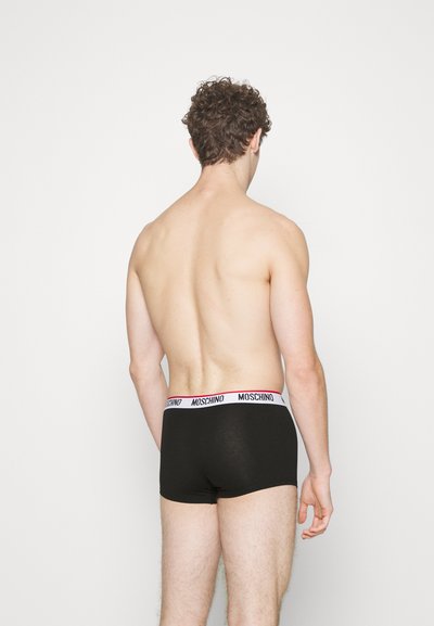Man seen from the back wearing black Moschino boxer briefs with a white waistband against a plain white background.