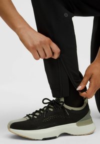 Juodos sportinės kelnės su užtrauktukais ant sijonų, derinamos su juodais ir alyvuogių spalvos sportbačiais, turinčiais tinklinį viršutinį sluoksnį ir paminkštintą pėdų dalį.