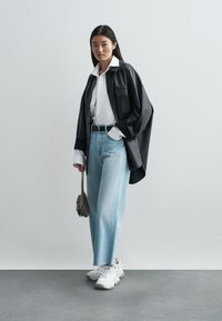 Chemise surdimensionnée en cuir noir, chemise blanche à col en dessous, jean bleu clair à jambes larges, baskets blanches et sac à main gris.
