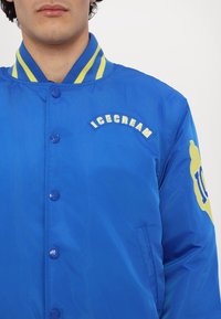 Chaqueta azul brillante con cierre de botones, acentos amarillos y "ICECREAM" bordado en blanco. Presenta un cuello deportivo a rayas y detalles de logo.