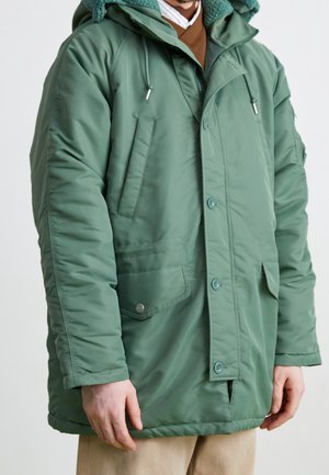 Personne portant une parka matelassée verte avec fermeture à boutons et fermeture éclair, poches avant et une capuche avec cordons.