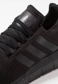 Svarta stickade sneakers med svarta snören, tre svarta ränder på sidan och en texturerad svart sula.