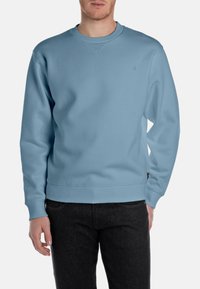 Felpa blu chiaro con scollo rotondo, polsini e orlo a coste, realizzata in tessuto morbido, con un piccolo logo sul petto. Indossata con jeans neri.