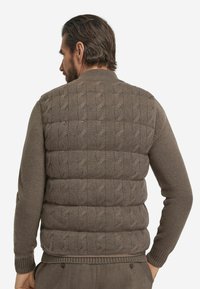 Braune gestrickte Weste mit Zopfmuster und Steppdesign. Gerippter Kragen und Saum. Das Material wirkt weich und strukturiert.