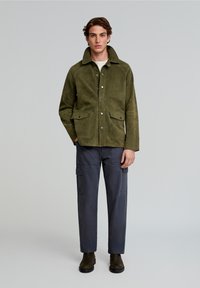 Olijfgroene suède jas met drukknoopjes, twee voorzakken, gecombineerd met marineblauwe cargobroek en donkerbruine enkelboots.