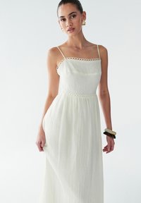 Witte geplooide maxijurk met spaghettibandjes, voorzien van een decoratieve kanten rand langs de halslijn en taille. Soepele, vloeiende stoftextuur.