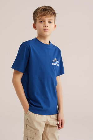 Băiat cu păr scurt purtând un tricou albastru "Puerto Montaña" și pantaloni cargo bej, stând cu o mână în buzunar pe un fundal neutru.