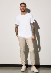 Bílý bavlněný tričko, béžové slim-fit chino kalhoty a světle zbarvené tenisky. Tetování viditelné na předloktí. Neutrální pozadí s jemnými stíny.