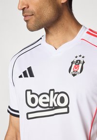 Camisola desportiva branca com detalhes em preto e vermelho. Apresenta riscas pretas da Adidas, um decote redondo e o logótipo "Beko" em destaque.