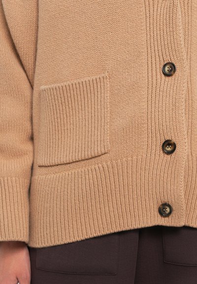 Beige kötött kardigán bordázott mintával, nagy gombokkal és egy rakott zsebbel az elején.