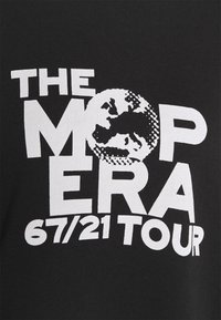 Camiseta negra con texto en blanco en negrita: "THE MOPERA" con un diseño de globo y "67/21 TOUR" en la parte inferior. 95% algodón para una textura suave.