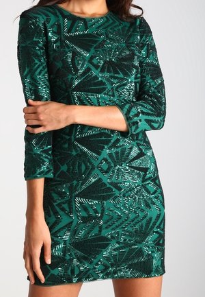 Kobieta ubrana w dopasowaną, zieloną sukienkę z długim rękawem ozdobioną czarnymi geometrycznymi wzorami z cekinów, z ramionami skrzyżowanymi na piersi i po boku.