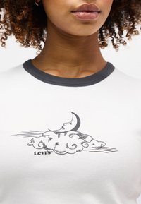 Κοντινό πλάνο ατόμου που φοράει λευκό πουκάμισο με μαύρο γιακά, με μαύρο γραφικό σχέδιο ημισελήνου με πρόσωπο πάνω από στροβιλιζόμενα σύννεφα και κείμενο "Levi's".