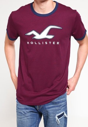 Bordowy bawełniany t-shirt z granatowym kołnierzem i mankietami, z białym logo ptaka i napisem „HOLLISTER” poniżej, widok z przodu, noszony z postrzępionymi dżinsami.