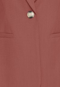 Rust-colored fabric coat with a single large translucent button and two horizontal slit pockets near the waist.