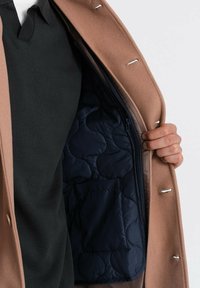 Abrigo de lana marrón con herrajes plateados, con un forro de acolchado azul marino y bolsillos. Camiseta de punto negra visible debajo.