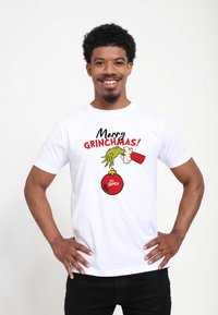 Bílý bavlněný tričko s nápisem "Veselé GRINCHMAS!" a grafikou ruky Grinče, která drží červenou ozdobu s nápisem "Grinch".