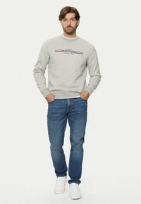 Sudadera gris con cuello redondo y logo "HILFIGER", combinada con jeans azules y zapatillas blancas. Corte casual y tejido texturizado.
