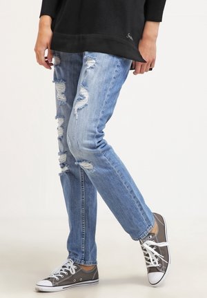 Personne portant un jean déchiré bleu clair et des baskets basses noires, debout avec une jambe légèrement pliée.