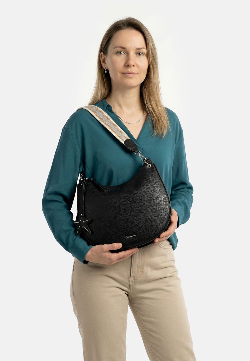 Femme en blouse sarcelle et pantalon beige tenant un sac à main en cuir noir avec une bandoulière large à rayures et un pendentif en forme d'étoile.