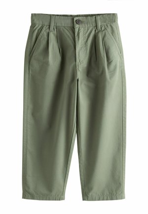 Băiat cu păr blond stă cu o mână în buzunar, purtând un tricou alb, pantaloni verde măsliniu și adidași albi pe fundal bej.